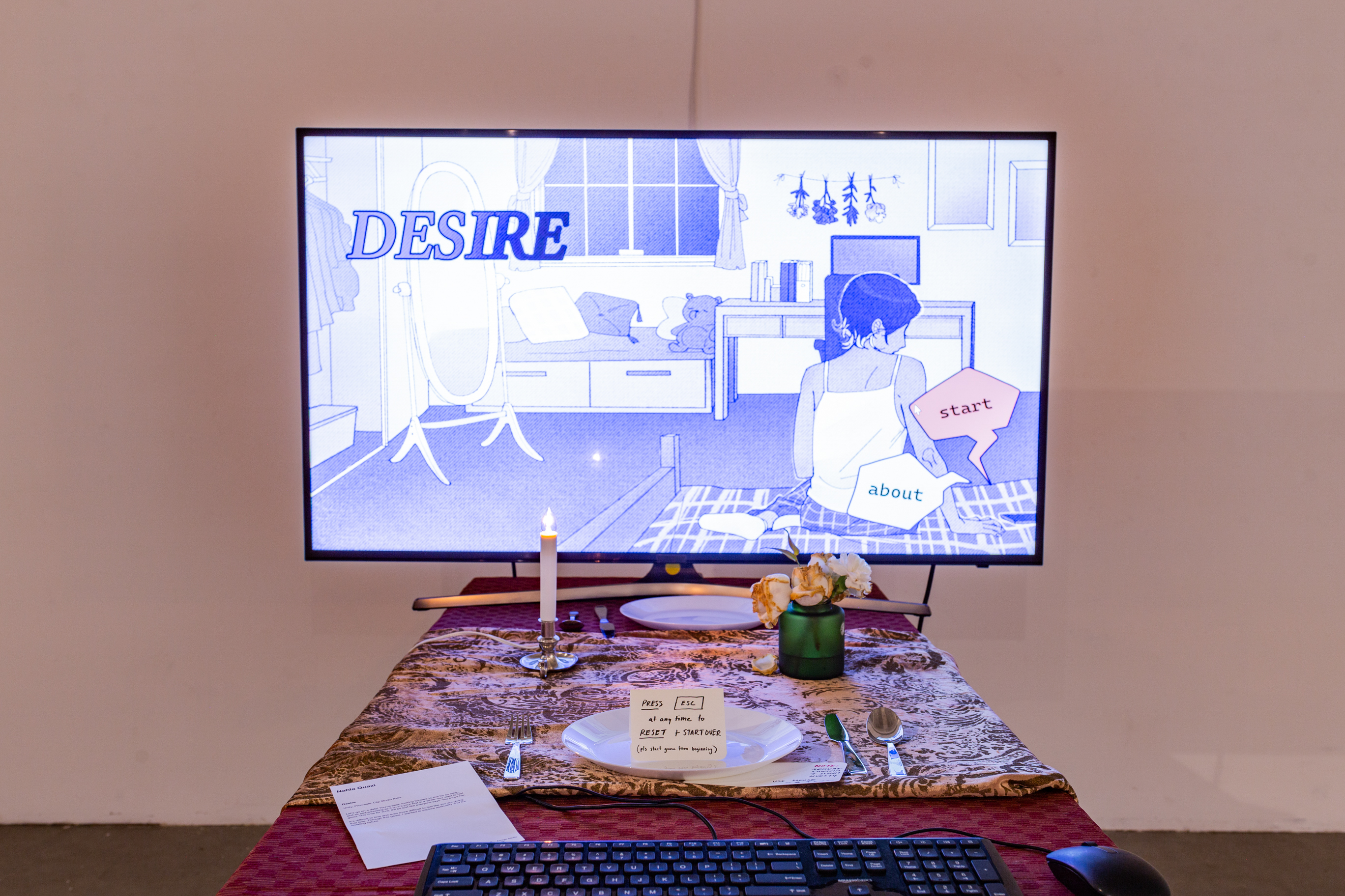 An upholstered chair sits in front of a table with tablecloth. On top of the table is a keyboard, a computer mouse, another table runner, a glass case with two flowers, a fake wax candle, and a television. On the TV is the start screen of an interactive game.